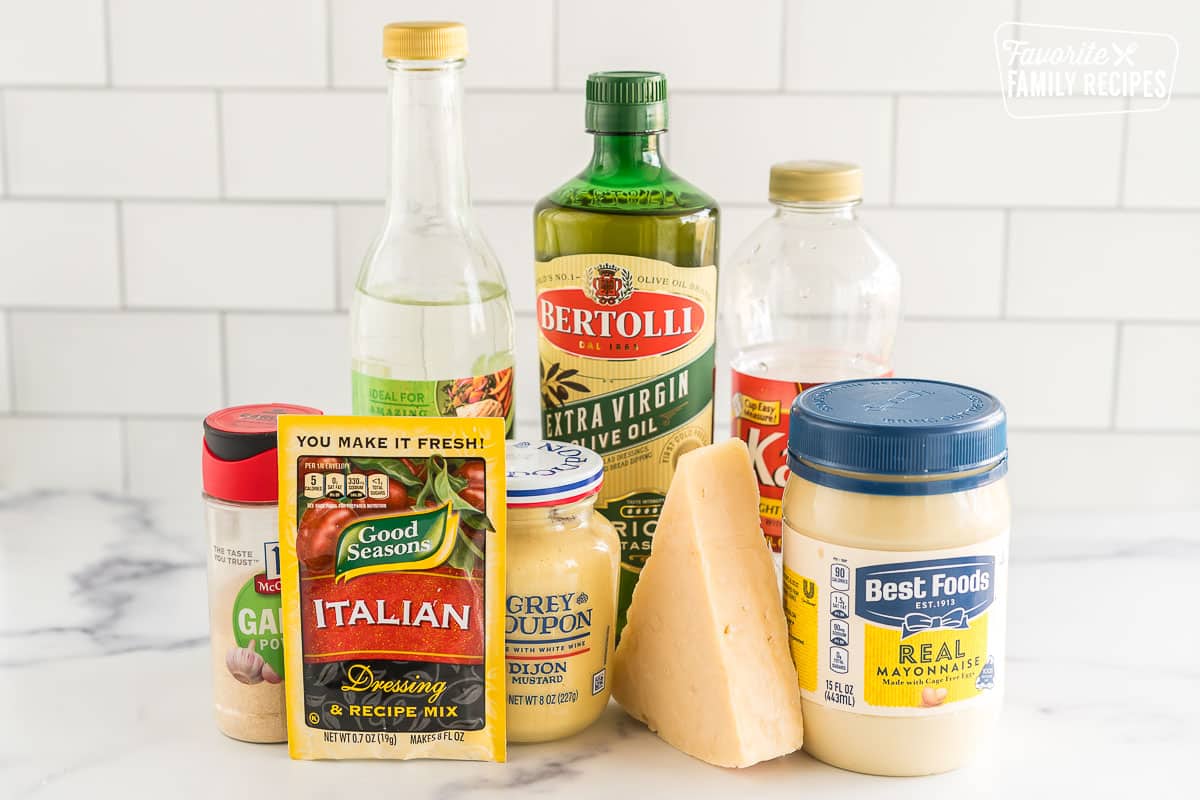 Olive oil, vinegar, corn syrup, garlic powder, Italian seasoning, mustard, parmesan, and mayo on a counter.