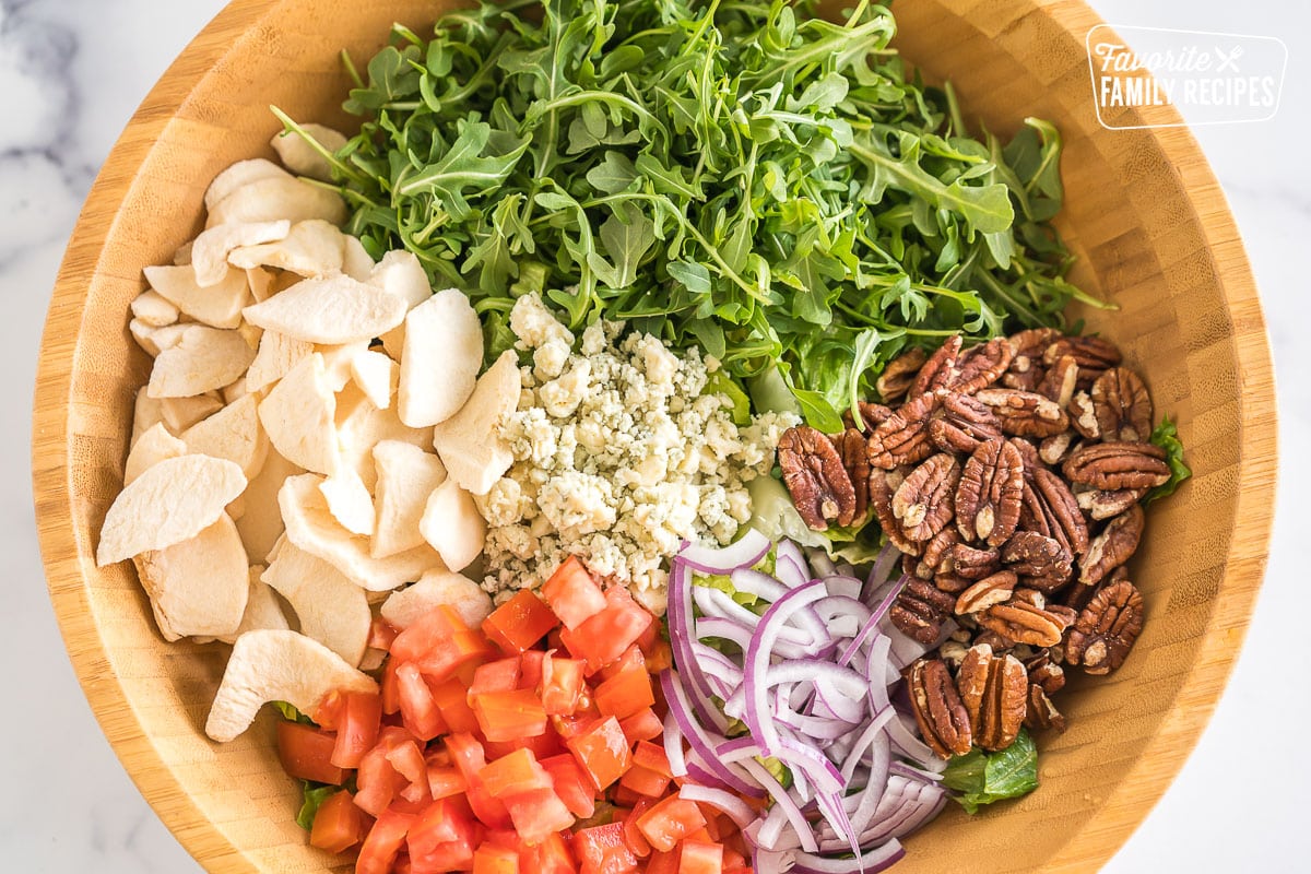 A large bowl with romaine, arugula, tomatoes, freeze-dried fruit, red onions, gorgonzola, and pecans