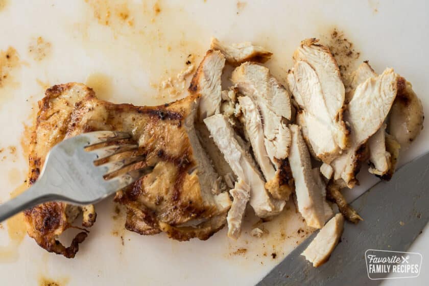 Sliced grilled chicken on a cutting board with a fork and knife