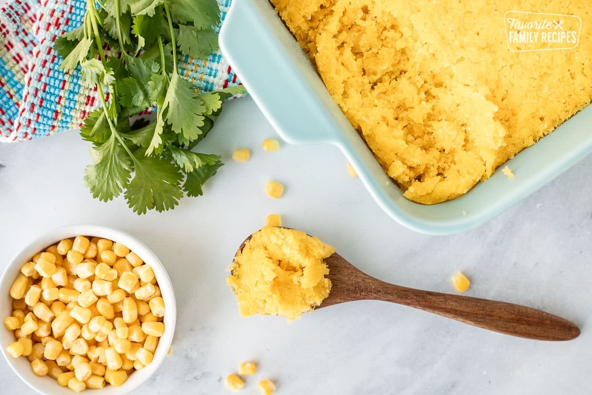 Spoon of Sweet Corn Cakes (Tomalitos) resting next to casserole dish of baked Sweet Corn Cakes (Tomalitos).