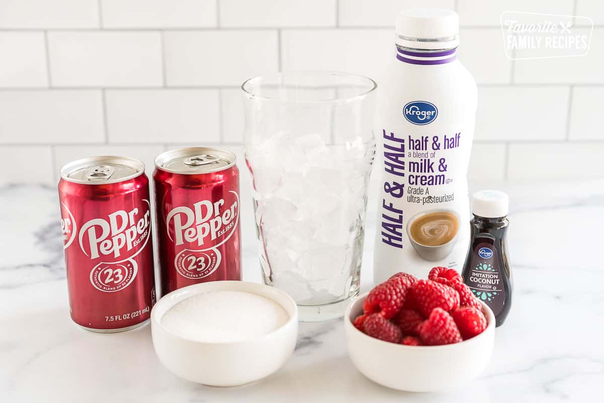 cans of Dr. Pepper, a cup half full of ice, half and half, coconut extract, raspberries, and sugar on the counter