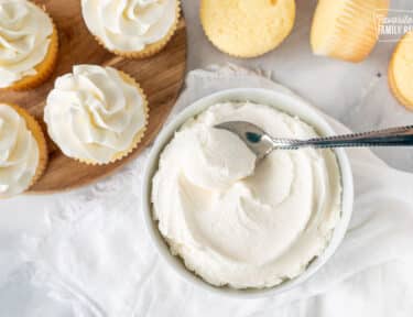 Bowl of Vanilla Buttercream Frosting with a spoon. Vanilla cupcakes on the side.