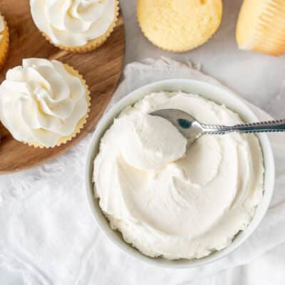 Bowl of Vanilla Buttercream Frosting with a spoon. Vanilla cupcakes on the side.