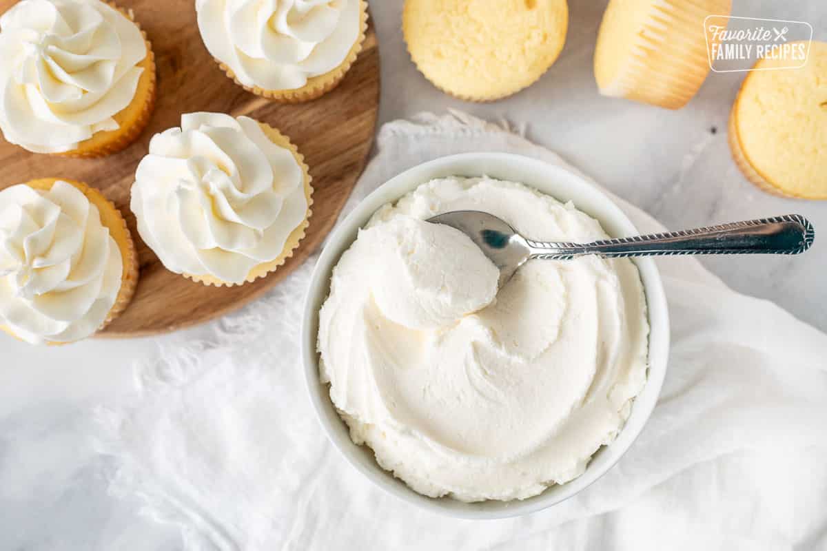 Bowl of Vanilla Buttercream Frosting with a spoon. Vanilla cupcakes on the side.