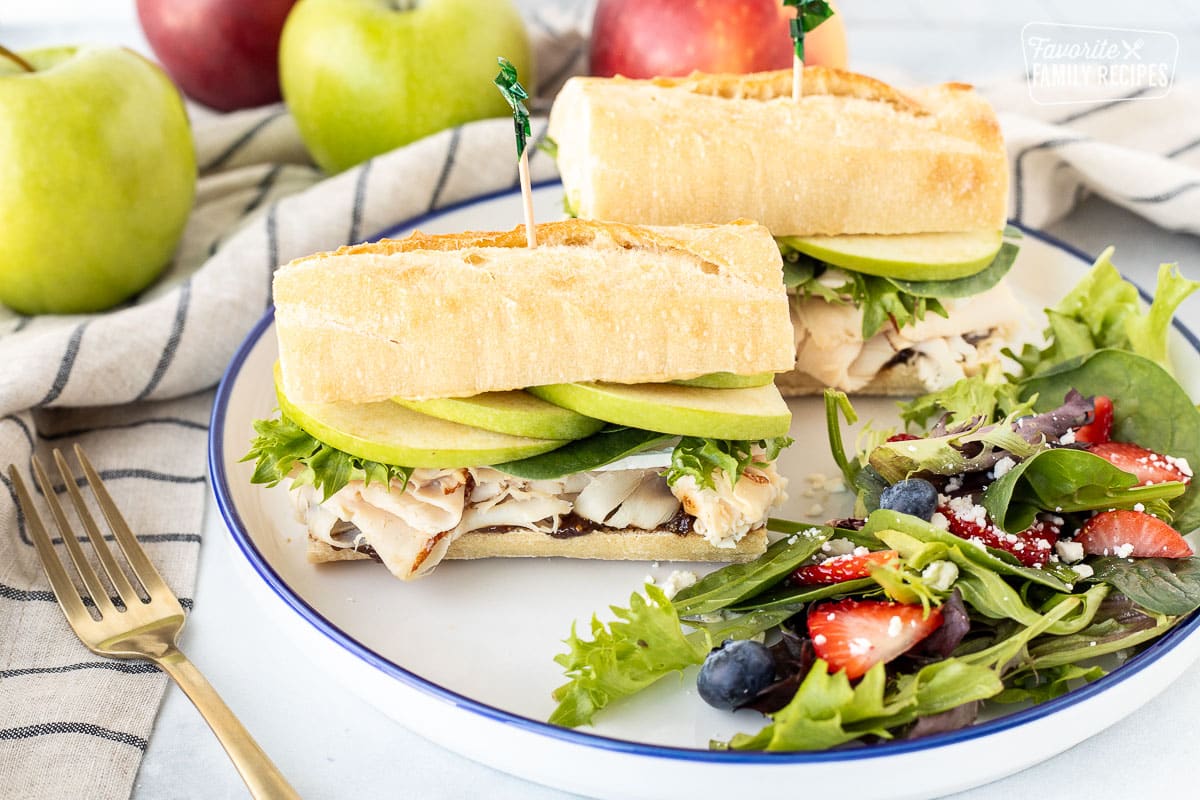 Two halves of a Turkey Sandwich with Brie Cheese and Apples on a plate with a side salad.
