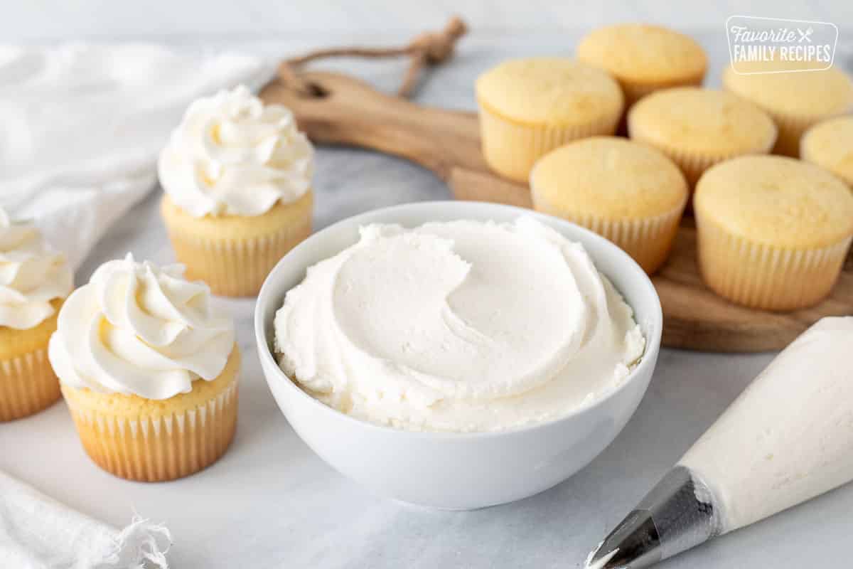 Bowl of Vanilla Buttercream Frosting next to a piping bag and vanilla cupcakes.