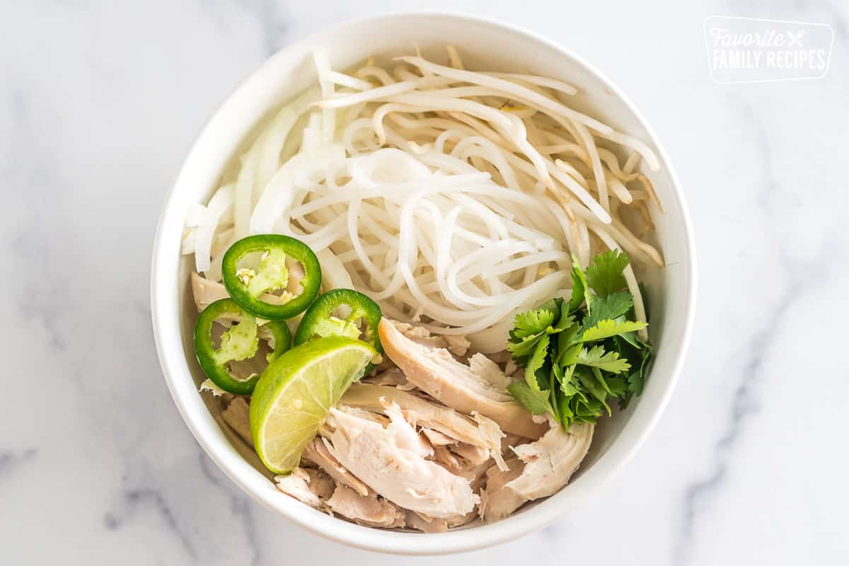 A bowl with rice noodles, bean sprouts, jalapeno slices, lime wedges, and chicken.