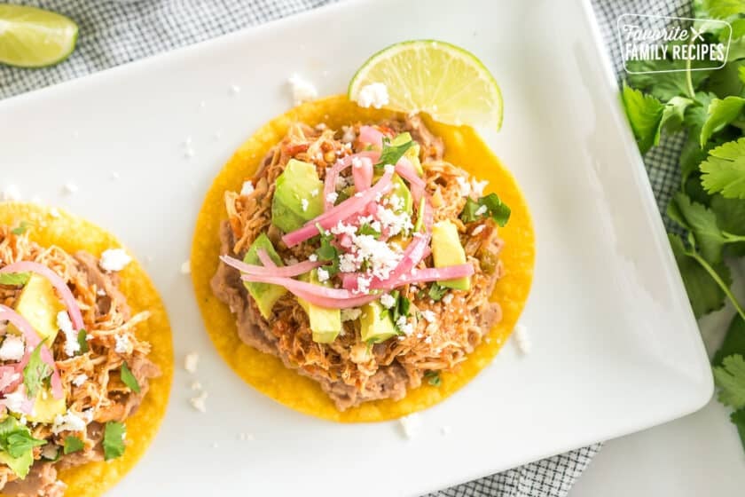 A corn tortilla with beans, shredded meat, avocado, cilantro, pickled red onion, and cotija cheese with a lime wedge