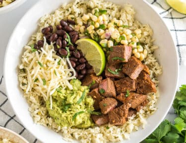 A bowl with rice, beans, cheese, steak, lime, guacamole, and corn salsa