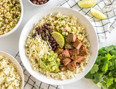 A bowl with rice, beans, cheese, steak, lime, guacamole, and corn salsa