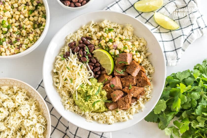 A bowl with rice, beans, cheese, steak, lime, guacamole, and corn salsa