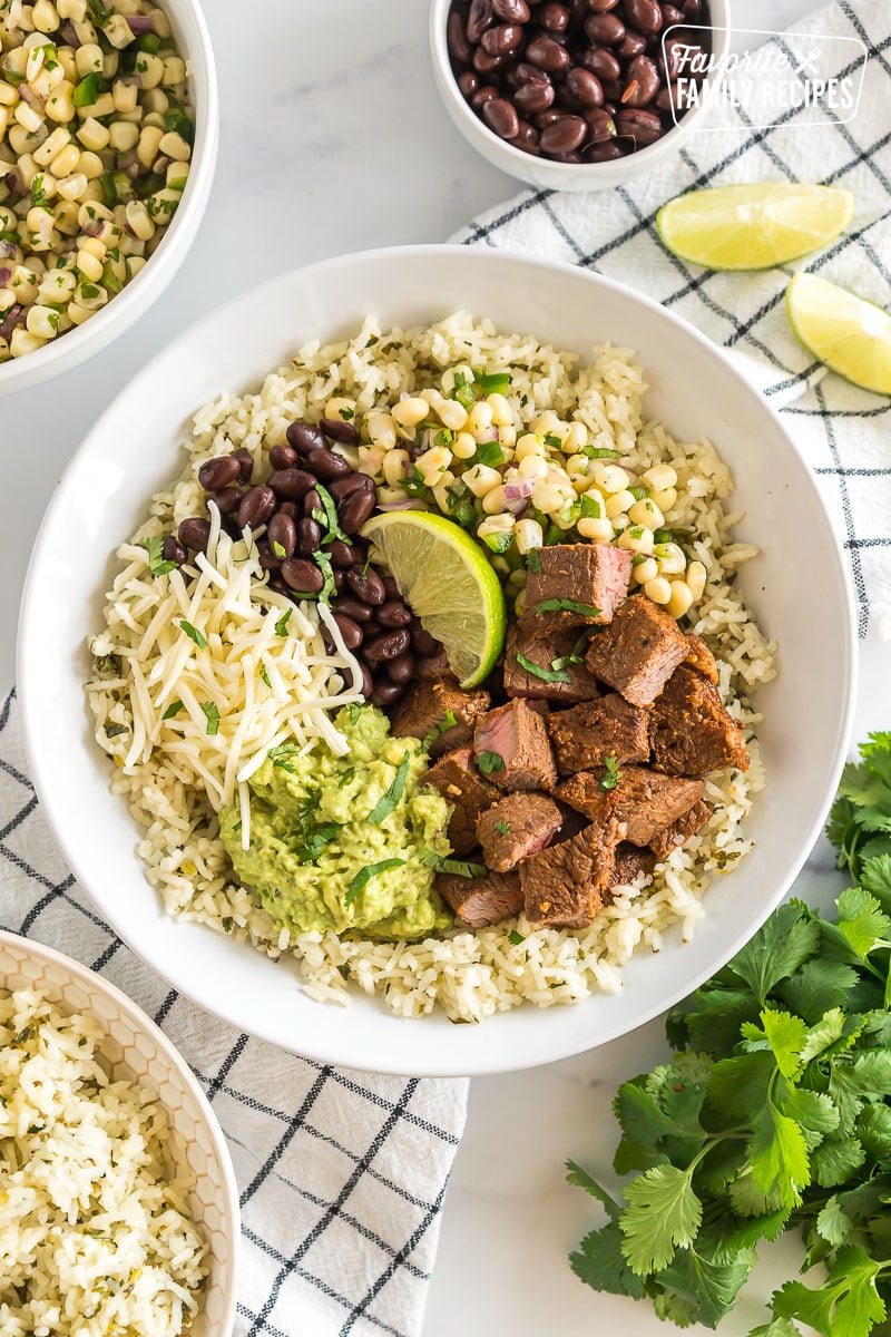 A bowl with rice, beans, cheese, steak, lime, guacamole, and corn salsa