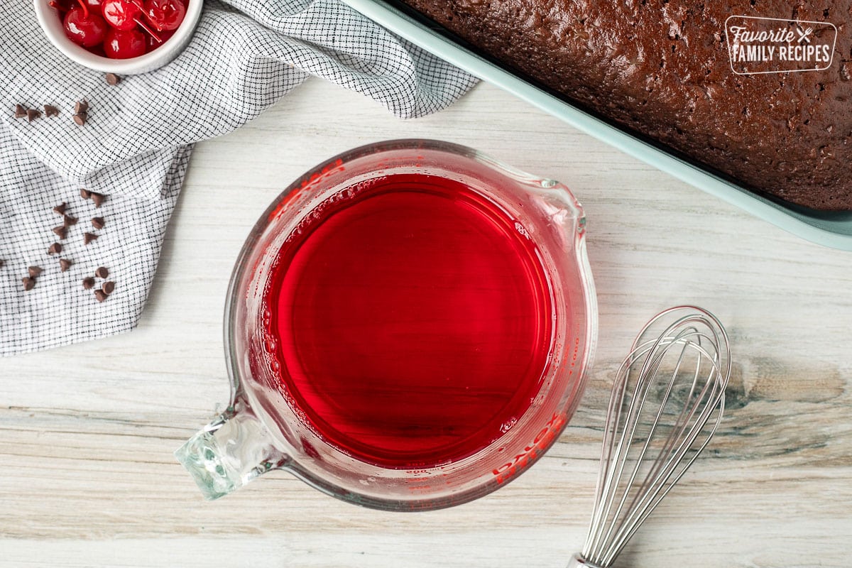 Bowl of liquid cherry jello next to a whisk and baked chocolate cake on the side.