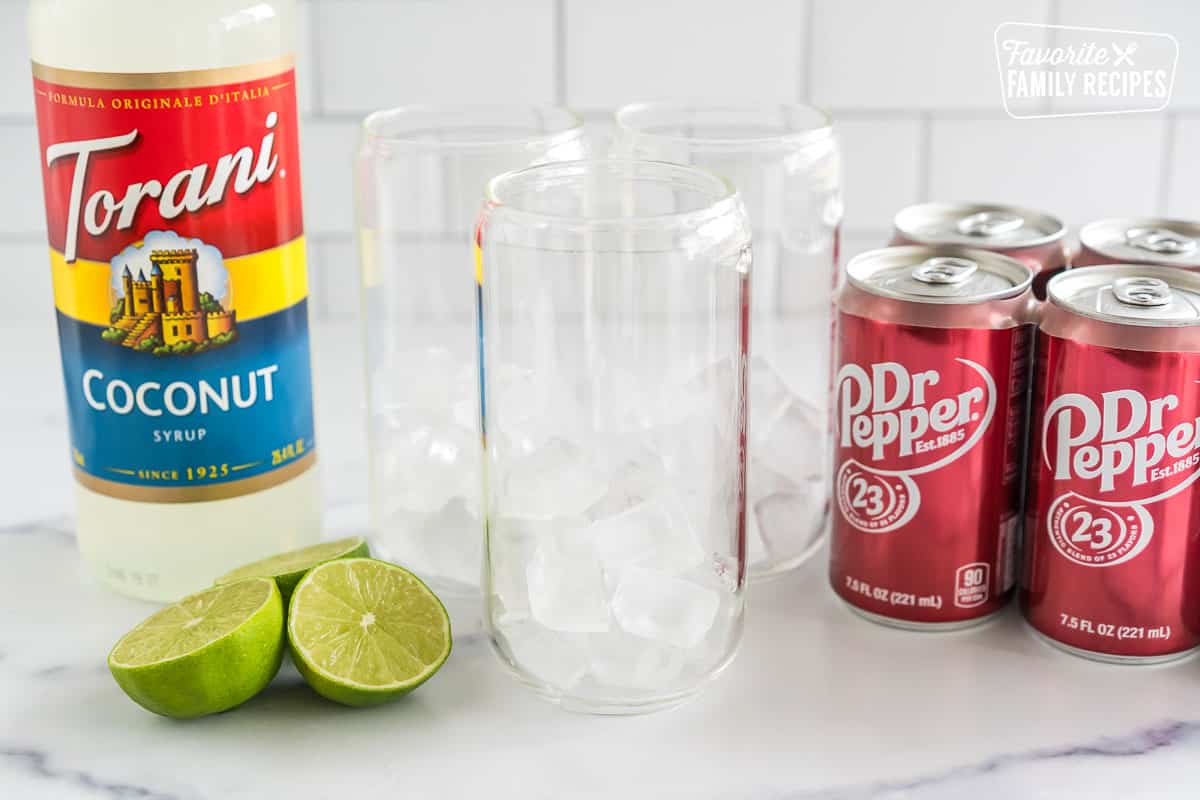 Cups with ice, cans of Dr Pepper, limes, and coconut syrup on the counter