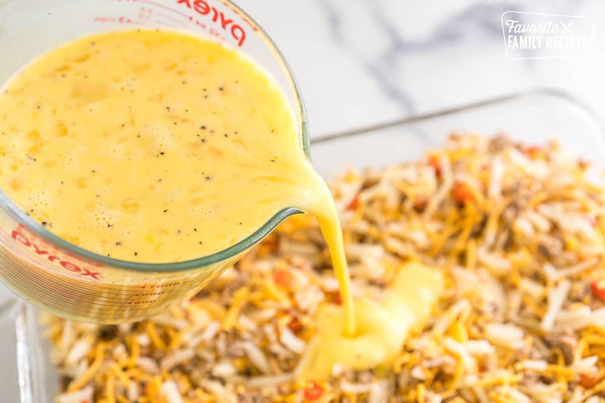 A glass measuring cup pouring egg mixture over a baking dish of hash browns, cheddar cheese, sausage, and veggies