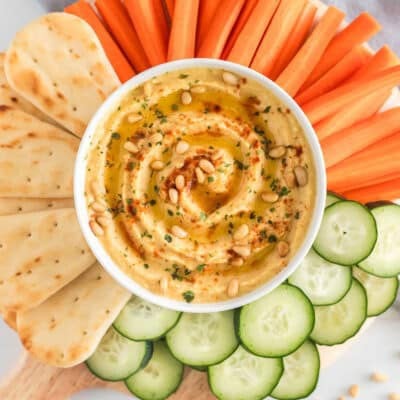 Homemade hummus topped with olive oil, pine nuts and paprika on a platter with naan bread, carrots and cucumbers