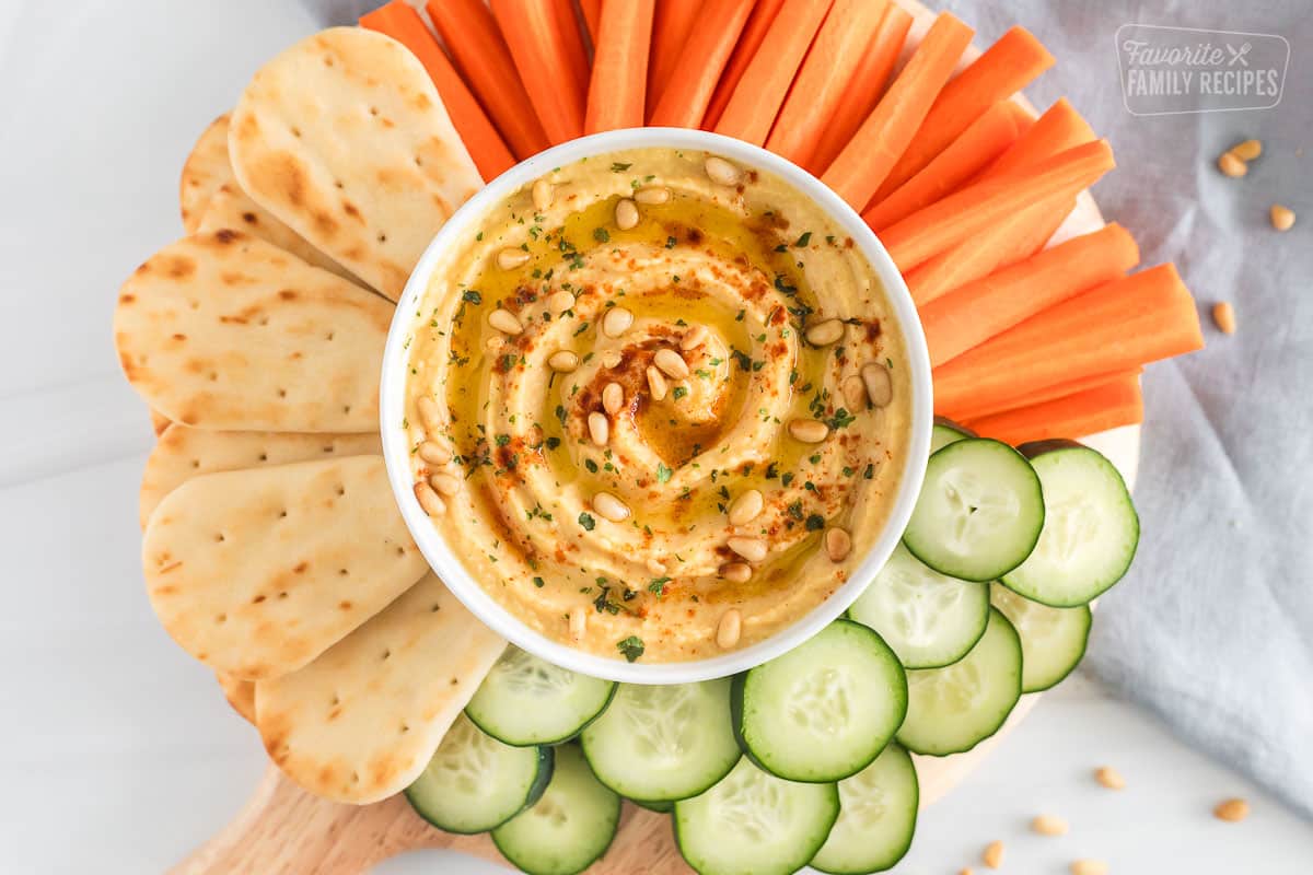 Homemade hummus topped with olive oil, pine nuts and paprika on a platter with naan bread, carrots and cucumbers