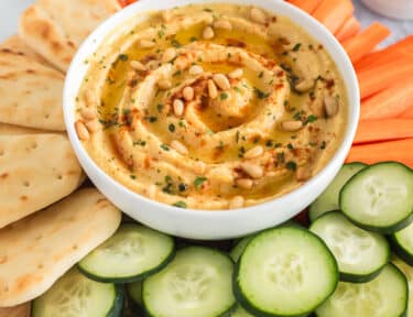 Homemade hummus topped with olive oil, pine nuts and paprika on a platter with naan bread, carrots and cucumbers