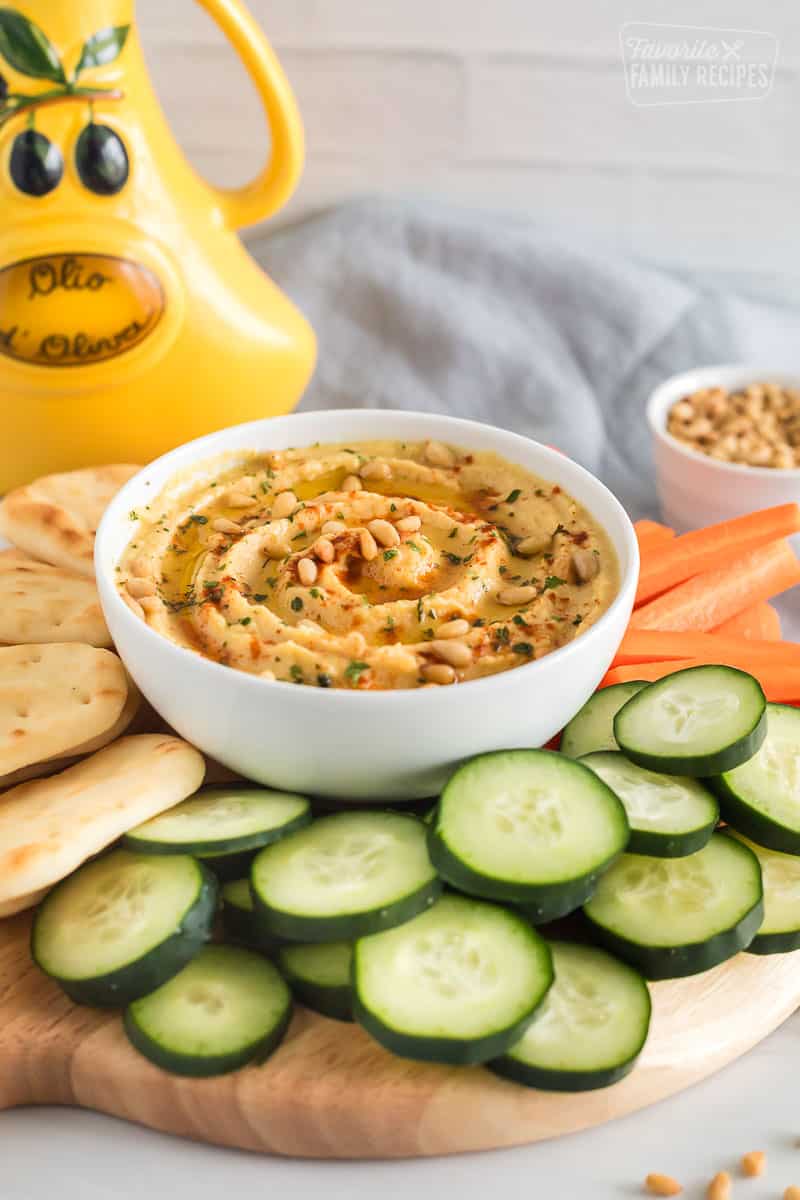 Homemade hummus topped with olive oil, pine nuts and paprika on a platter with naan bread, carrots and cucumbers