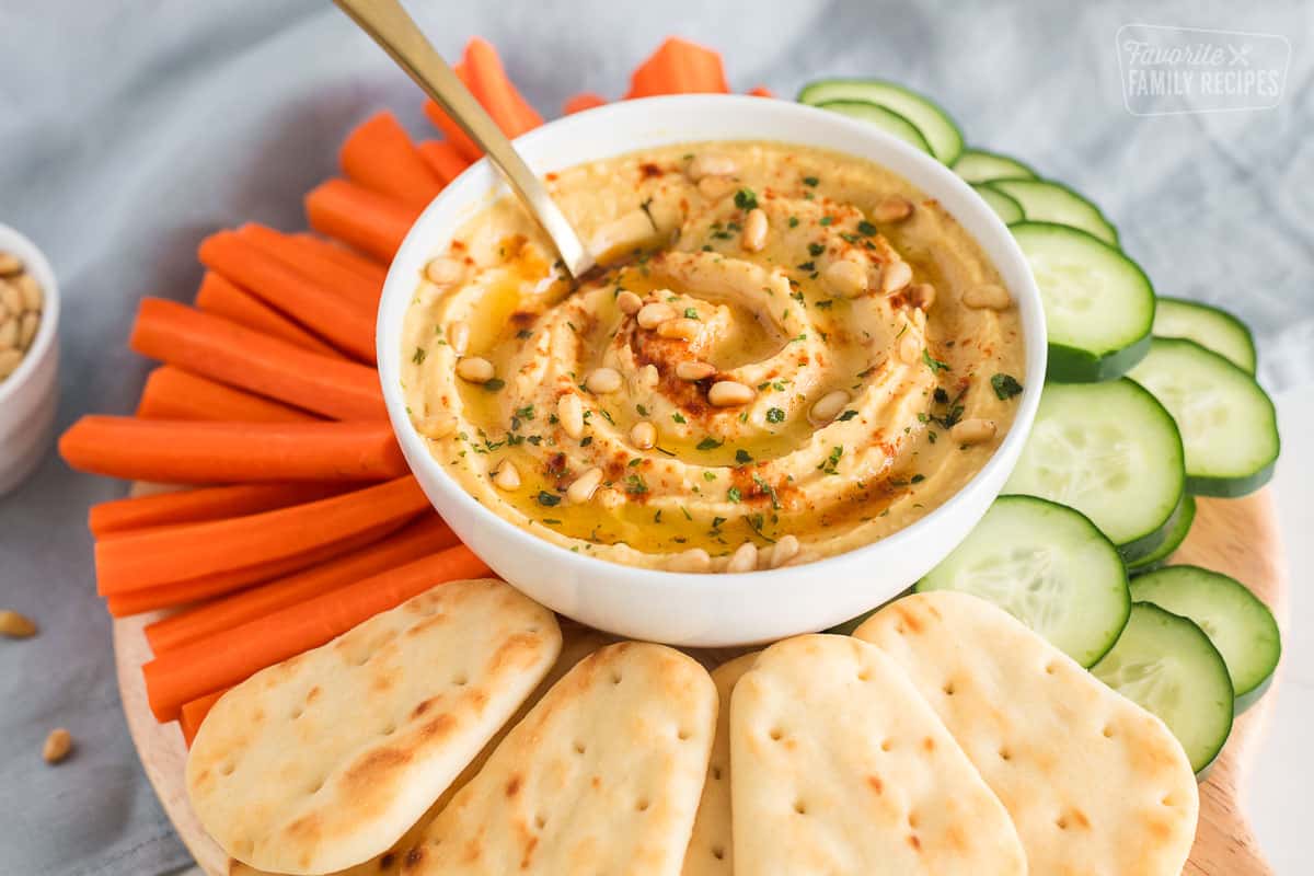 Homemade hummus topped with olive oil, pine nuts and paprika on a platter with naan bread, carrots and cucumbers