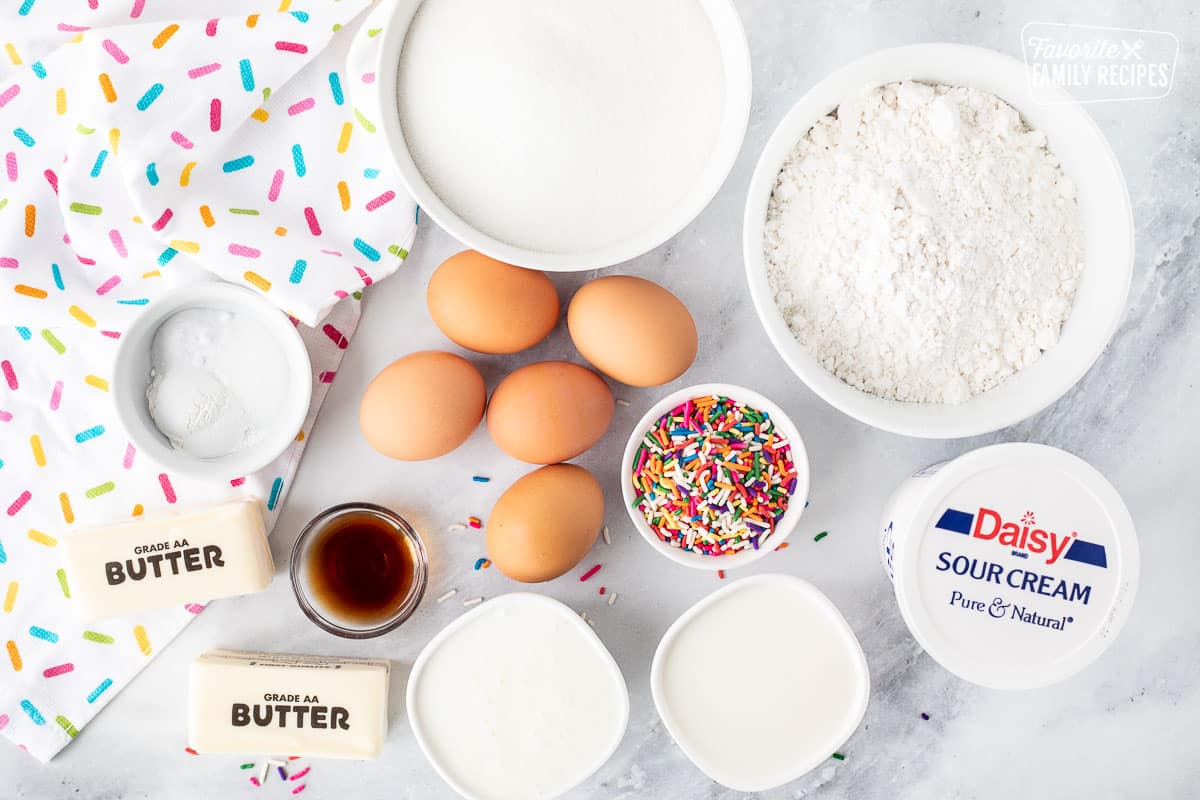 Ingredients to make Funfetti cake recipe including sugar, cake flour, sour cream, sprinkles, eggs, butter, milk, buttermilk, vanilla, baking soda, baking powder and salt.