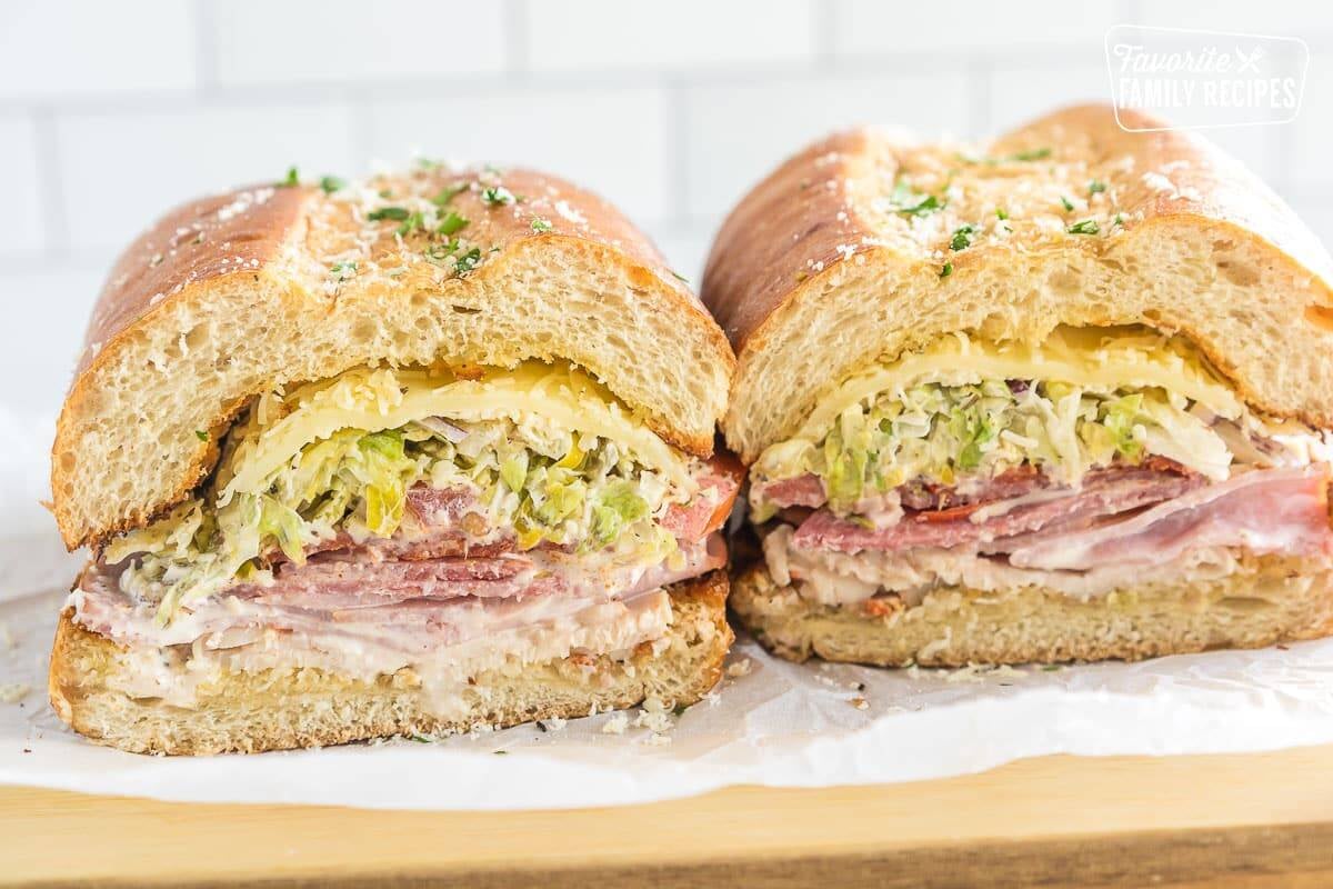 A Grinder sandwich cut in half on a cutting board, showing a cross section of layers of meats, cheese, and salad.