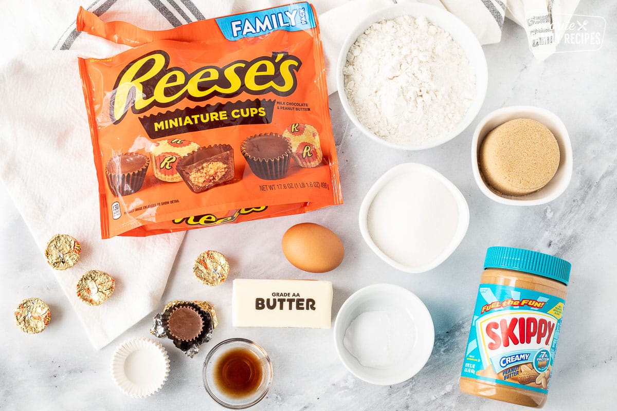 Ingredients to make Reese's Peanut Butter Cup Cookies including Reese's miniature cups, flour, brown sugar, sugar, peanut butter, salt, baking soda, butter, egg, vanilla and mini muffin liners.