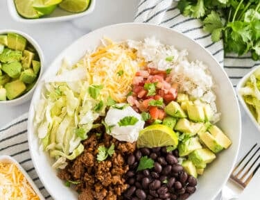 A Taco Bowl with rice, beans, taco meat, cheese, lettuce, avocado, cilantro, and lime