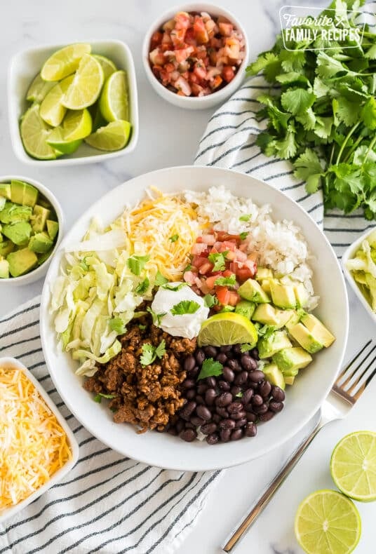 A Taco Bowl with rice, beans, taco meat, cheese, lettuce, avocado, cilantro, and lime