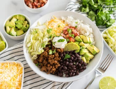 A Taco Bowl with rice, beans, taco meat, cheese, lettuce, avocado, cilantro, and lime