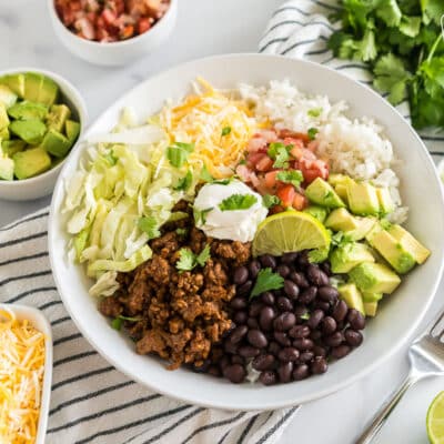 A Taco Bowl with rice, beans, taco meat, cheese, lettuce, avocado, cilantro, and lime