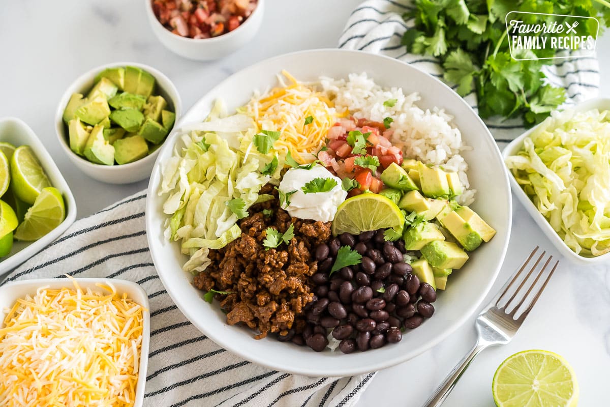 A Taco Bowl with rice, beans, taco meat, cheese, lettuce, avocado, cilantro, and lime