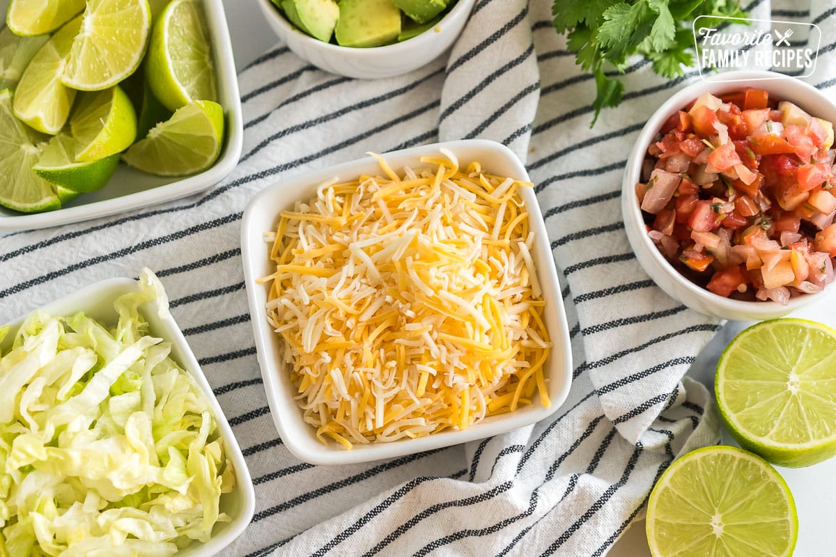 little bowls of cheese, pico de gallo, lettuce, limes, avocado, lime, and cilantro.