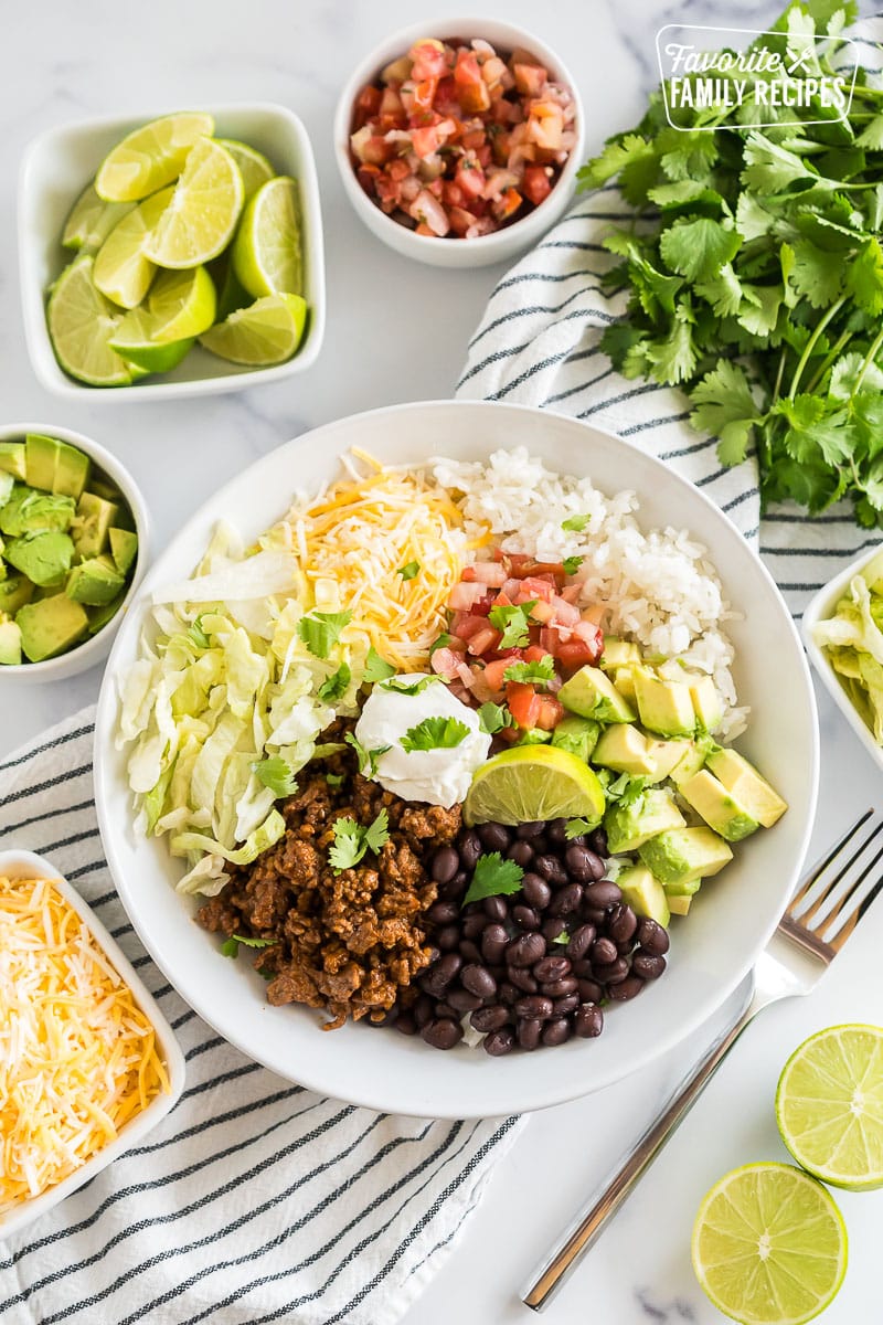 A Taco Bowl with rice, beans, taco meat, cheese, lettuce, avocado, cilantro, and lime.