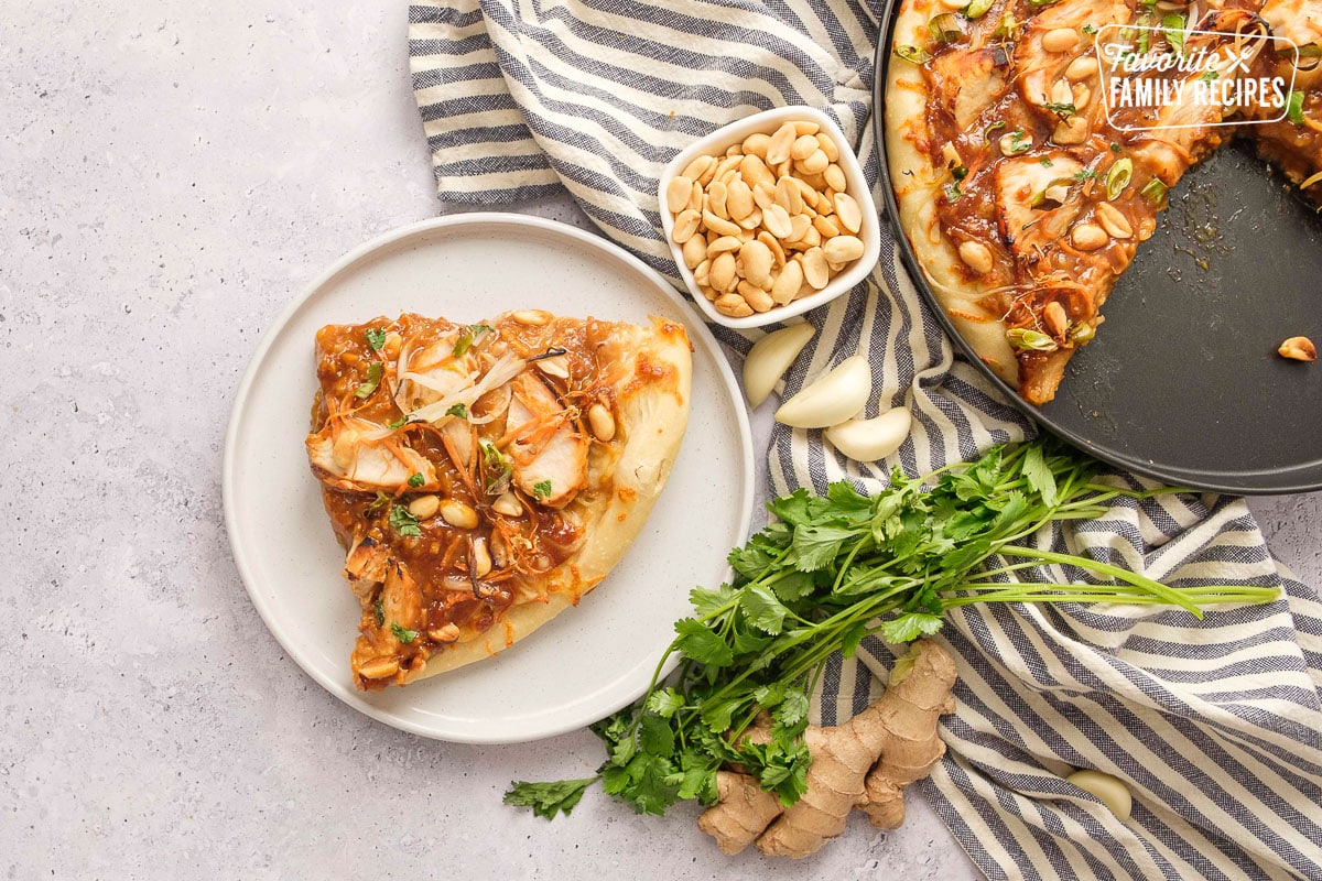 A slice of Thai chicken pizza on a white plate with peanuts in a side bowl and the whole pizza on the side. 