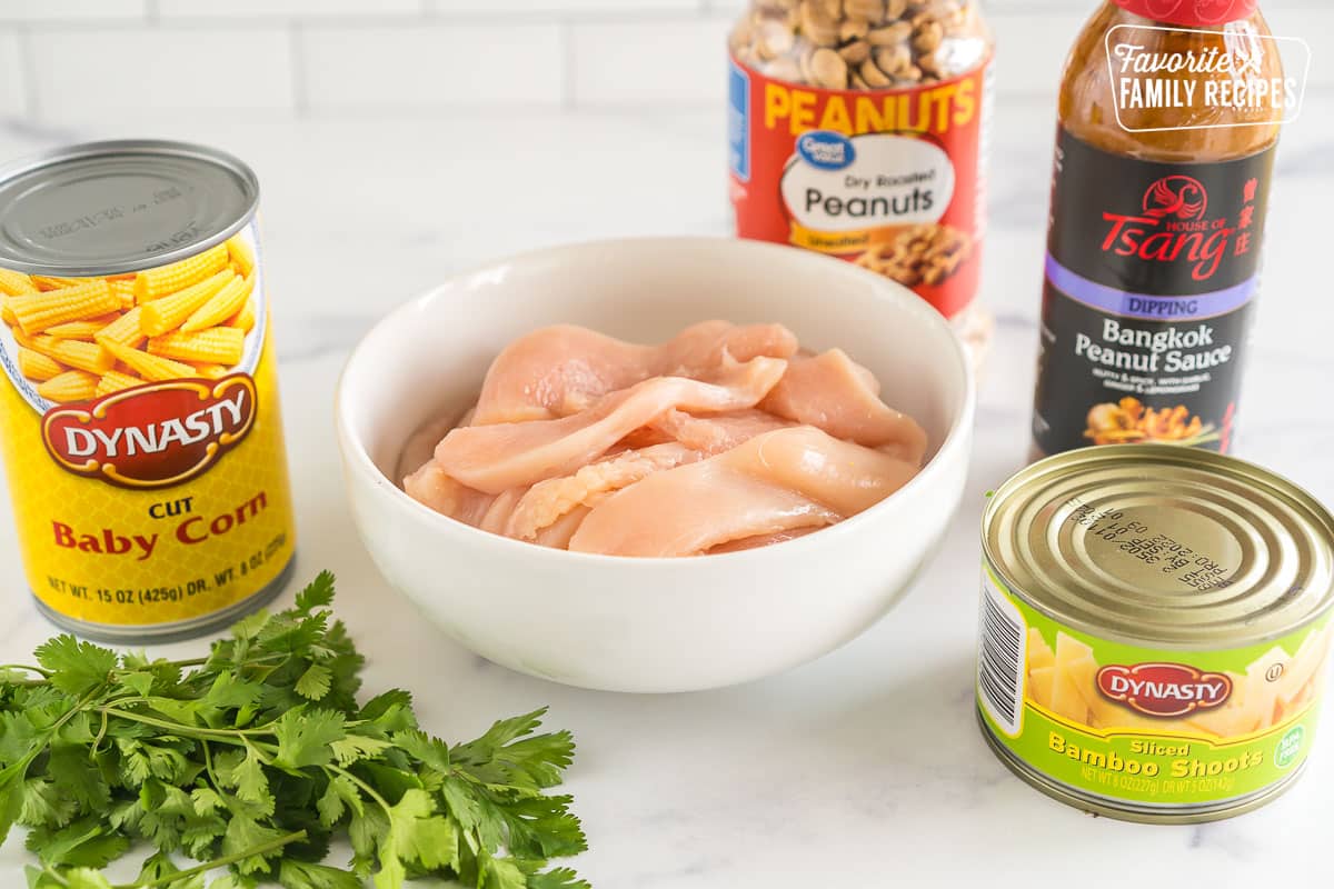 A bowl of sliced raw chicken, a can of baby corn, peanuts, peanut sauce, a can of bamboo shoots, and cilantro on the counter top