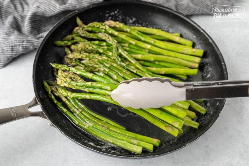 Tongs holding Sweet Sautéed Asparagus over a skillet.