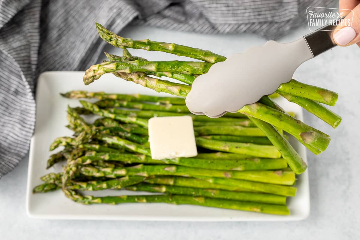 Tongs holding up Sweet Sautéed Asparagus over a plate of Sweet Sautéed Asparagus.