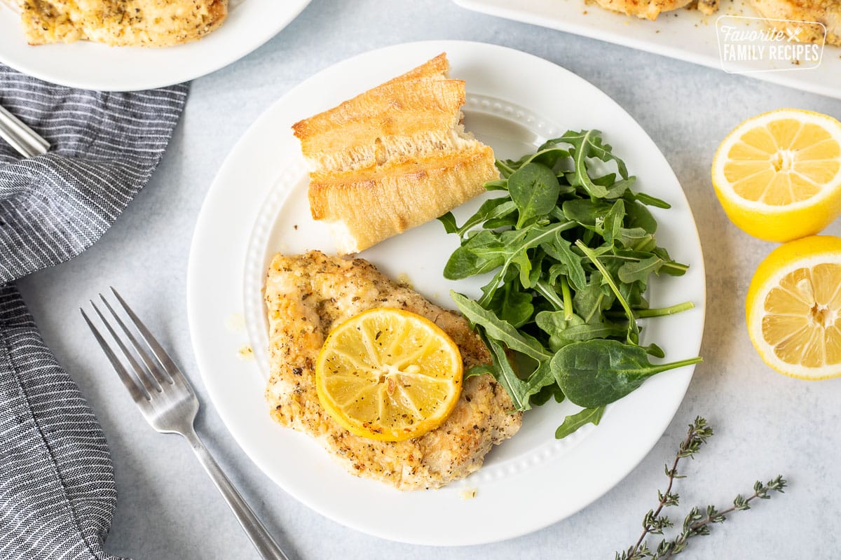 Plate with Baked Lemon Chicken breast with sliced lemon, fresh salad greens and a piece of French bread.