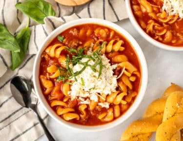 Bowl of Lasagna soup with cheeses mix on top with fresh cut basil. Bread sticks on the side.