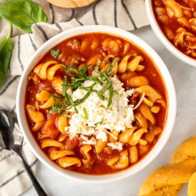 Bowl of Lasagna soup with cheeses mix on top with fresh cut basil. Bread sticks on the side.