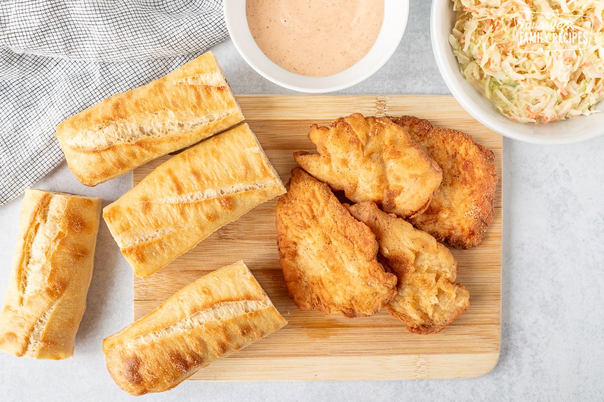 Baguette pieces and fried chicken breasts on a cutting board. Bowl of coleslaw and dressing on the side.