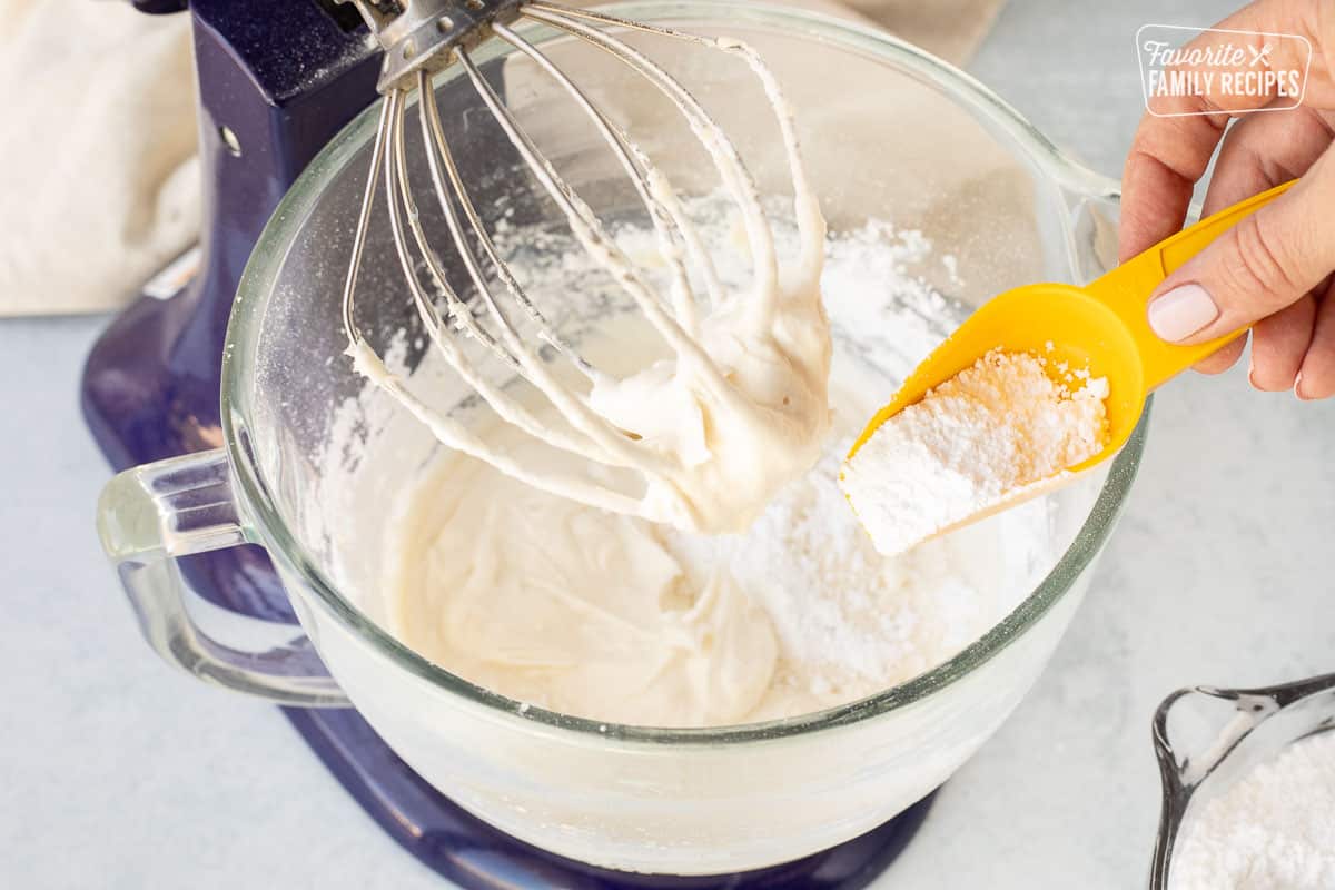 Pouring powdered sugar into a glass mixing bowl of cream cheese and butter frosting.