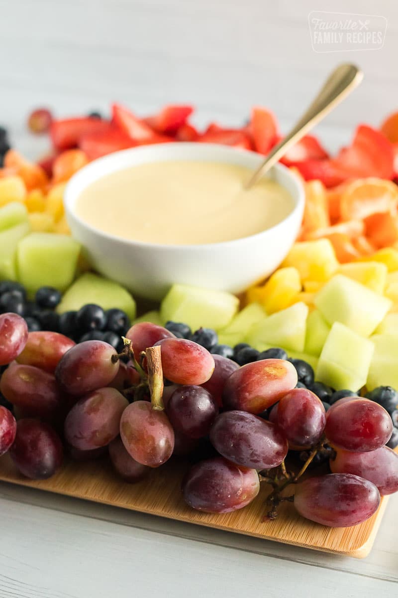 Cream cheese fruit dip with a spoon on a platter with strawberries, orange slices, pineapple, honeydew, blueberries, and grapes.