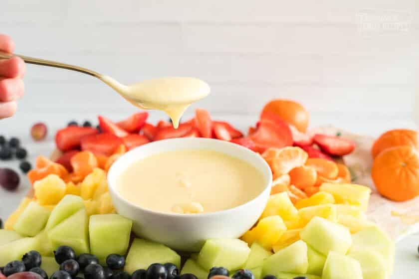 Cream cheese fruit dip with a spoon on a platter with strawberries, orange slices, pineapple, honeydew, blueberries, and grapes