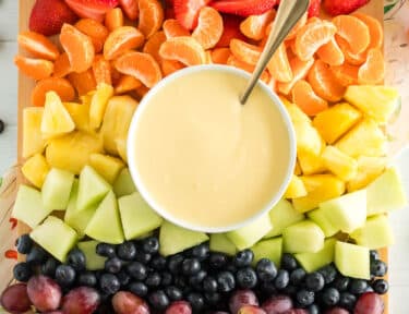 Cream cheese fruit dip with a spoon on a platter with strawberries, orange slices, pineapple, honeydew, blueberries, and grapes