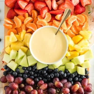 Cream cheese fruit dip with a spoon on a platter with strawberries, orange slices, pineapple, honeydew, blueberries, and grapes