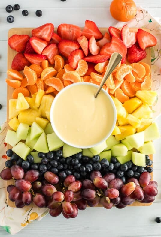 Cream cheese fruit dip with a spoon on a platter with strawberries, orange slices, pineapple, honeydew, blueberries, and grapes