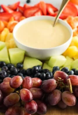 Cream cheese fruit dip with a spoon on a platter with strawberries, orange slices, pineapple, honeydew, blueberries, and grapes
