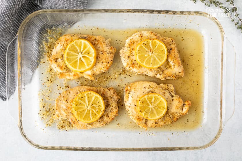 Baking dish with four baked lemon chicken breasts with seasoning on top and baked lemon slices on top.